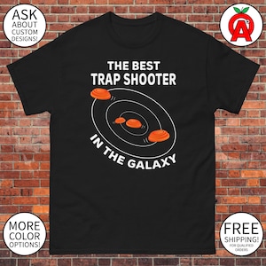 May include: Black t-shirt with white text that reads "The Best Trap Shooter In The Galaxy". The text is surrounded by a circle with three orange clay pigeons flying around it.