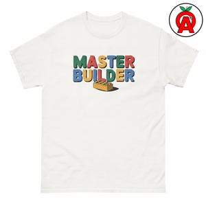 Könnte beinhalten: Ein weißes kurzärmeliges T-Shirt mit dem Text MASTER BUILDER in mehrfarbigen Blockbuchstaben. Unter dem Text befindet sich eine kleine Abbildung eines einzelnen gelben Kunststoff-Bausteins mit einem darunter liegenden Schatten.