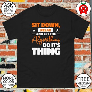 May include: Black t-shirt with the text "SIT DOWN, RELAX - AND LET THE Algorithms DO IT'S THING" in orange and white. The shirt has a classic fit and short sleeves. Additional text includes "ASK ABOUT CUSTOM DESIGNS!" and "MORE COLOR OPTIONS!"