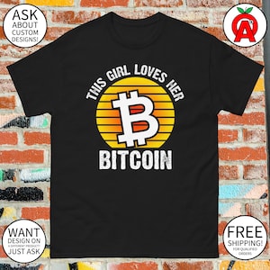 May include: Black t-shirt with a white graphic of a bitcoin symbol in a sunset. The text "This girl loves her Bitcoin" is written around the symbol.