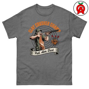 Puede incluir: Camiseta gris con un gráfico de una persona disparando una escopeta a un plato de arcilla. El texto "CLAY CRUSHER CHAMP" está encima de la figura, con "Pull. Aim. Dust." en una pancarta debajo. El diseño utiliza naranja, marrón y verde.
