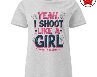 Puede incluir: Camiseta gris claro con el texto "YEAH, I SHOOT LIKE A GIRL + WANT A LESSON?" en rosa y azul oscuro. La camiseta tiene cuello redondo y mangas cortas. Un logotipo está en la esquina superior derecha.