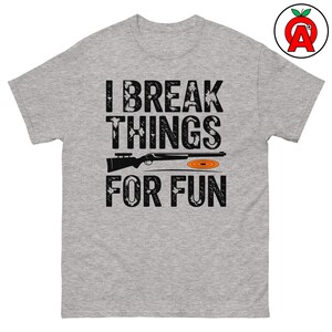 Könnte beinhalten: Ein graues T-Shirt mit dem Text "I break things for fun" in Schwarz. Unter dem Text befindet sich eine Schwarzweißillustration einer Flinte und einer Tontaubenscheibe.