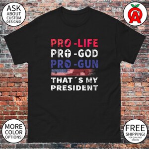 Pode incluir: Camiseta preta com um gráfico desbotado em branco, vermelho e azul que diz "Pro-Life, Pro-God, Pro-Gun, That's My President".