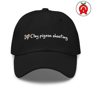 May include: Black baseball cap with the embroidered text "I ❤️ Clay pigeon shooting" in white. The heart symbol is red. The cap has a curved brim and a button on top. A logo with a red apple and the letter "A" is on the upper right.
