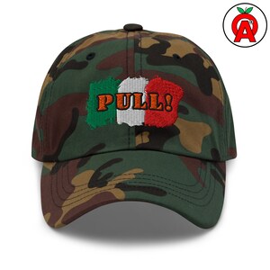 May include: Camouflage baseball cap with the word "PULL!" embroidered in orange over a design resembling the Italian flag. The cap is a mix of green, brown, and black.