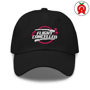 Könnte beinhalten: Schwarze Baseballkappe mit dem gestickten Text "FLIGHT CANCELLED" in Weiß, mit einem rosa Kreis und zwei weißen Gewehren. Die Kappe hat einen gebogenen Schirm und einen Knopf oben. Ein Logo mit einem roten Apfel und dem Buchstaben "A" befindet sich an der Seite.