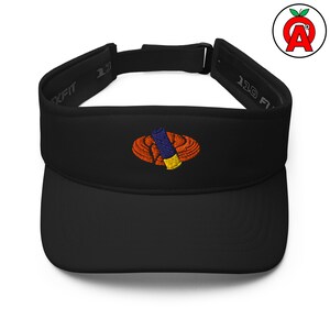 May include: Black visor with an embroidered design of a blue and yellow object inside an orange oval. The visor has a curved brim and an adjustable strap. The text "110 FIT" is visible on the side.