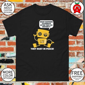 May include: Black t-shirt featuring a yellow robot illustration with the text "WHAT HAPPENS TO ROBOTS AFTER THEY GO DEFUNCT? THEY RUST IN PEACE!". The shirt also has the text "ASK ABOUT CUSTOM DESIGNS!" and "MORE COLOR OPTIONS!"