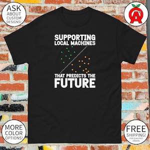 May include: Black t-shirt with white text: "SUPPORTING LOCAL MACHINES THAT PREDICTS THE FUTURE." The design includes green triangles and orange squares. Additional text includes "ASK ABOUT CUSTOM DESIGNS!" and "MORE COLOR OPTIONS!"