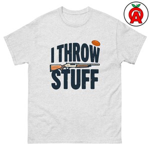 Puede incluir: Una camiseta gris claro con el texto "I THROW STUFF" en azul marino, con una escopeta y un plato de arcilla. La camiseta es de estilo cuello redondo clásico.