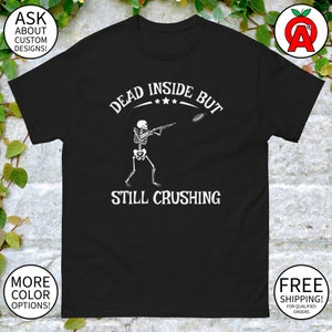 Puede incluir: Camiseta negra con un esqueleto blanco disparando a una paloma de arcilla. El texto "Dead Inside But Still Crushing" está impreso en la camiseta.