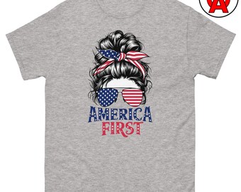 Puede incluir: Una camiseta gris jaspeado con un gráfico de un moño desordenado con una cinta roja, blanca y azul, y gafas de aviador con lentes de la bandera estadounidense. Debajo del gráfico, el texto dice AMERICA FIRST en letras azules y rojas.