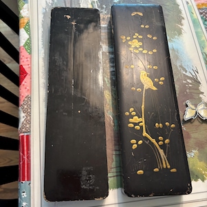 Puede incluir: Dos cajas rectangulares de laca negra. Una caja es de color negro liso con signos visibles de desgaste. La otra caja presenta un diseño pintado en oro de un pájaro posado sobre una rama florida. Ambas cajas tienen bordes redondeados.