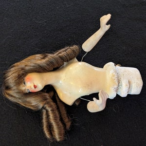 May include: A vintage porcelain doll torso with a brown wig and painted facial features. The doll's arms are detached and the torso is missing its legs.