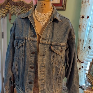 Puede incluir: Una chaqueta vaquera azul claro con un collar de perlas y una blusa de encaje blanco debajo. La chaqueta tiene cierre de botones y dos bolsillos en la parte delantera.