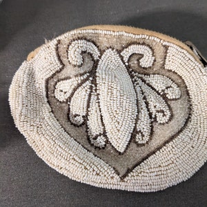 May include: A small, round, beige beaded purse with a white and brown floral design. The purse has a zipper closure.