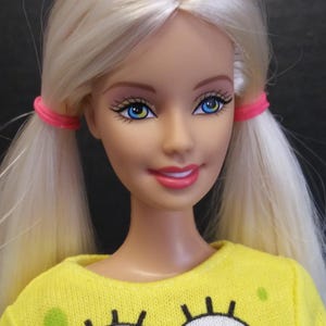 May include: A doll with long blonde hair styled in two pigtails secured with bright pink elastic bands. The doll has blue eyes, pink lipstick, and wears a yellow long-sleeved shirt featuring a large graphic of Spongebob Squarepants' eyes.