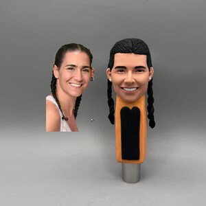 May include: A custom tap handle with a sculpted face, dark braided hair, and a smiling expression. The tap handle has an orange and black body. A photograph of the person is next to the tap handle. The base is silver.