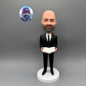 May include: A custom bobblehead figurine of a man in a black suit, holding an open book. The figure has a bald head and a beard. A small oval photo of the man wearing a beanie is in the upper left corner. The figurine stands on a white circular base.