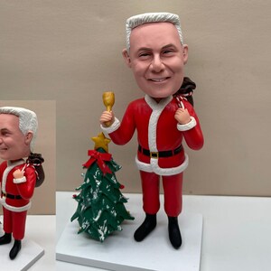 May include: A personalized Santa Claus figurine with a white beard and a red suit. The figurine is holding a bell in one hand and a bag of gifts in the other. The figurine is standing next to a small green Christmas tree.