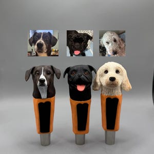Op de afbeelding: Drie op maat gemaakte biertapkranen met hondenportretten. De kranen zijn oranje met zwarte accenten en tonen een zwart-witte hond, een zwarte mops en een witte poedel.