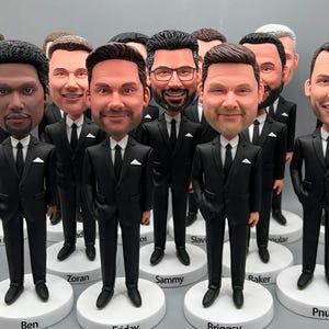 Könnte beinhalten: Eine Gruppe von 12 schwarzen und weißen Figuren von Männern in schwarzen Anzügen und weißen Hemden. Jede Figur hat ein anderes Gesicht und steht auf einem weißen Sockel mit einem aufgedruckten Namen.