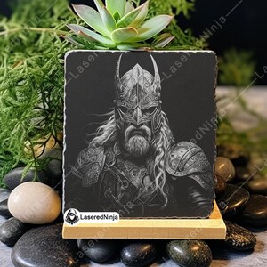 May include: Black and white engraved image of a Viking warrior wearing a horned helmet and armour. The image is on a black slate coaster with a wooden base.