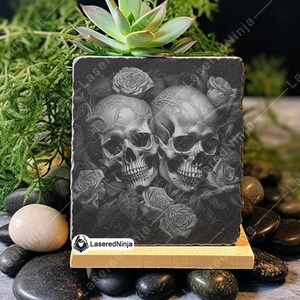 May include: Black and white engraved image of two skulls with roses on a black slate coaster. The coaster is on a wooden stand.