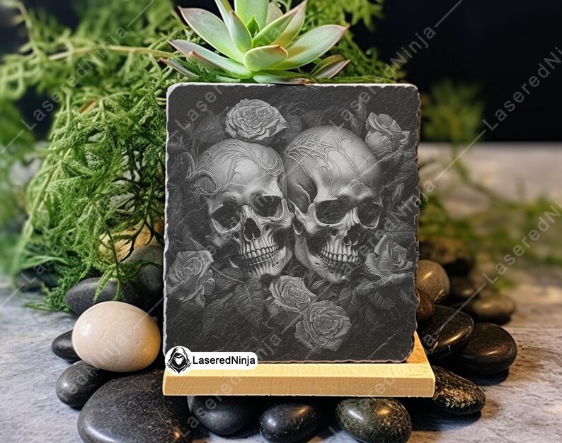 May include: Black and white engraved image of two skulls with roses on a black slate coaster. The coaster is on a wooden stand.