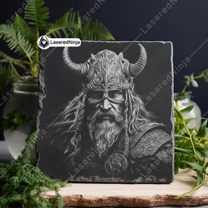 May include: Black and white engraved image of a Viking warrior with a horned helmet and long beard on a black slate coaster.