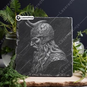 May include: A black slate coaster with a detailed engraving of a Viking warrior wearing a horned helmet. The engraving is in a silver color.