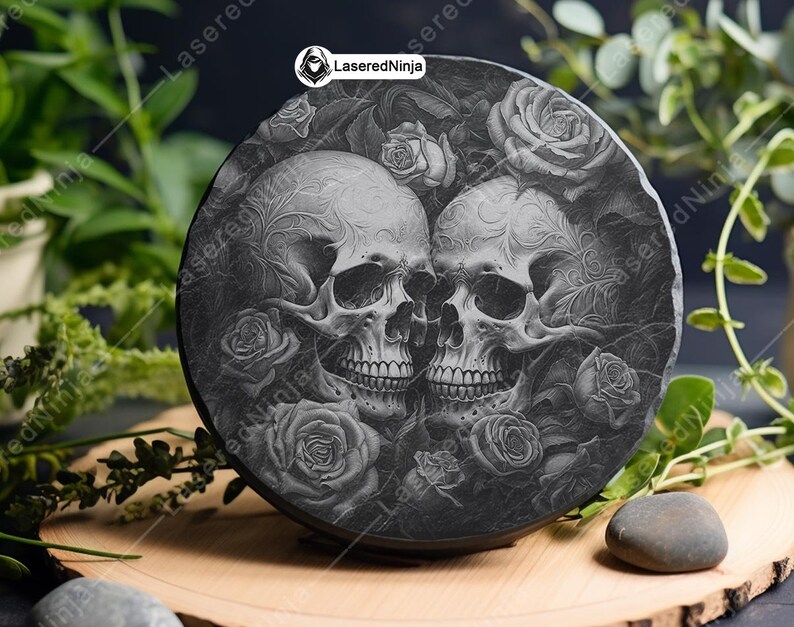 May include: A black slate coaster with a laser-etched design of two skulls with intricate details, surrounded by roses. The coaster is round and has a smooth, polished surface.
