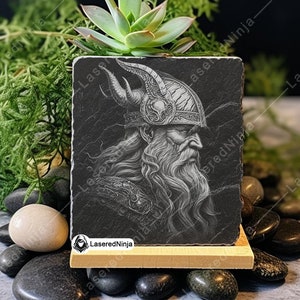 May include: A black and white engraved image of a Viking warrior with a horned helmet and a long beard. The image is on a black slate coaster with a wooden base.