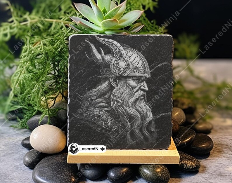 May include: A black and white engraved image of a Viking warrior with a horned helmet and a long beard. The image is on a black slate coaster with a wooden base.
