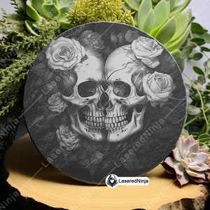 May include: A round black slate coaster with a design of two white skulls with roses around them. The skulls are facing each other and have a detailed design. The roses are white and have a realistic look. The coaster is on a wooden surface.