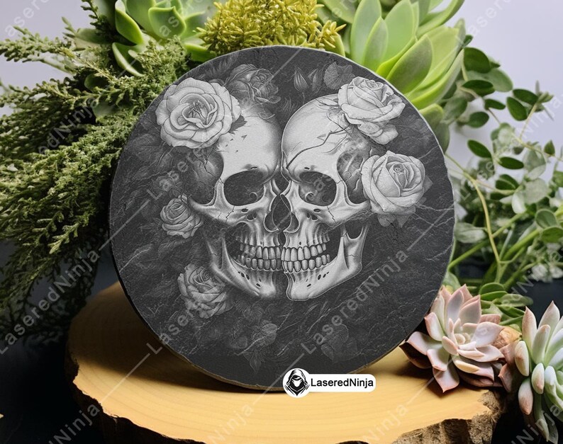 May include: A round black slate coaster with a design of two white skulls with roses around them. The skulls are facing each other and have a detailed design. The roses are white and have a realistic look. The coaster is on a wooden surface.