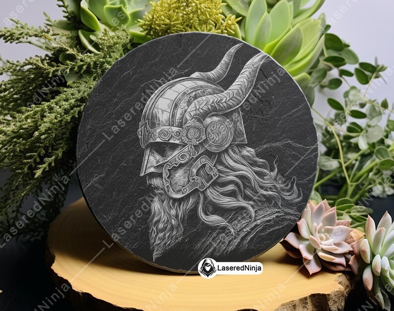 May include: A black slate coaster with a detailed engraving of a Viking warrior wearing a horned helmet. The engraving is in a white color and the background is black.