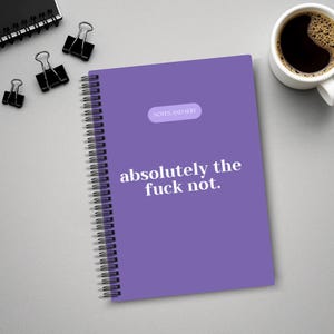 May include: A purple spiral-bound notebook with the text "absolutely the fuck not." and "NOTES AND SHIT" printed on the cover. The notebook is on a gray surface, with black binder clips and a cup of coffee.