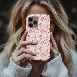 May include: Pink phone case featuring a repeating pattern of bubble tea cups with red bows, hearts, and striped straws. The case has a glossy finish and is designed to fit a smartphone. The overall design is cute and playful.