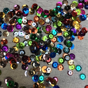 May include: A large assortment of colourful, iridescent sequins in various shades of blue, green, red, purple, and gold. The sequins are scattered on a light grey surface.