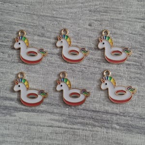 May include: Six metal charms shaped like white unicorn pool floats with gold-tone outlines. Each charm features a rainbow-colored mane and tail, a small gold horn, and a red base. A small circular loop is attached to the top of each unicorn head.