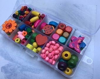 May include: A clear plastic storage container filled with assorted wooden beads in various shapes and colors. The collection includes flower, ladybug, heart, and butterfly shapes, alongside round, square, and cylindrical beads in shades of pink, yellow, green, blue, red, and purple.