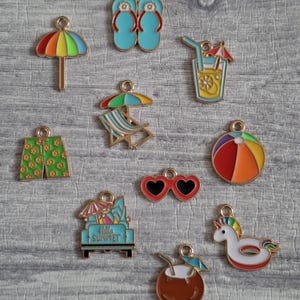 Peut inclure: Collection de breloques colorées sur le thème de l'été. Les breloques comprennent un parapluie arc-en-ciel, des tongs bleues, un cocktail, une chaise de plage, des lunettes de soleil en forme de cœur, un ballon de plage, un short, une voiture avec le texte "Hello Summer", une boisson à la noix de coco et un flotteur licorne.