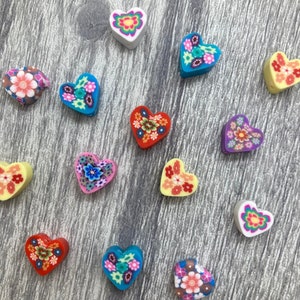 以下が含まれることがあります： 花柄のデザインが施されたカラフルなハート型のポリマークレイチャーム14個の詰め合わせ。各チャームには、赤、青、黄、ピンク、紫など、さまざまな色の異なる花柄が施されています。