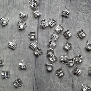 May include: A group of silver metal bead caps scattered on a grey wooden surface. The caps have a cylindrical shape with a small loop at the top.