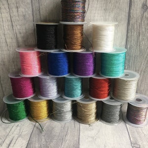 Puede incluir: Una colección de 20 carretes de hilo metálico en varios colores, incluyendo dorado, plateado, rojo, azul, verde y rosa. Los carretes están dispuestos en forma de pirámide sobre una superficie de madera.