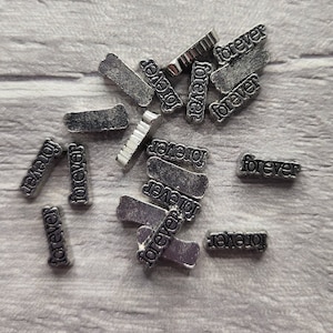 Può includere: Una collezione di ciondoli in metallo argentato, ognuno con la parola "forever" incisa. I ciondoli sono sparsi su una superficie di legno bianco.