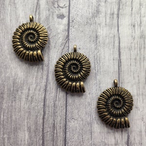 May include: Three antique gold-toned metal ammonite shell charms. The charms are detailed with a spiral pattern and have a loop at the top for attaching to jewellery or other crafts.