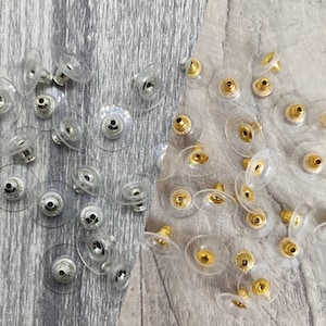 May include: A close-up of two groups of earring backs. The group on the left has silver posts and the group on the right has gold posts. Both groups are made of clear plastic.